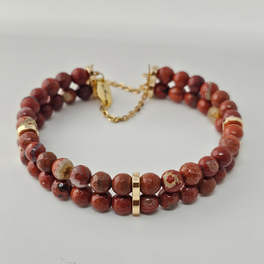 A double-strand bracelet made of faceted 4mm Red Jasper beads, featuring rich earthy red tones with natural variations. Accented with gold-tone spacers, a clasp, and a delicate gold chain with an engraved tag.