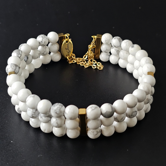 Triple strand bracelet made with 6mm round white howlite beads featuring subtle gray veining, accented with gold-tone spacers and chain clasp on a black background.