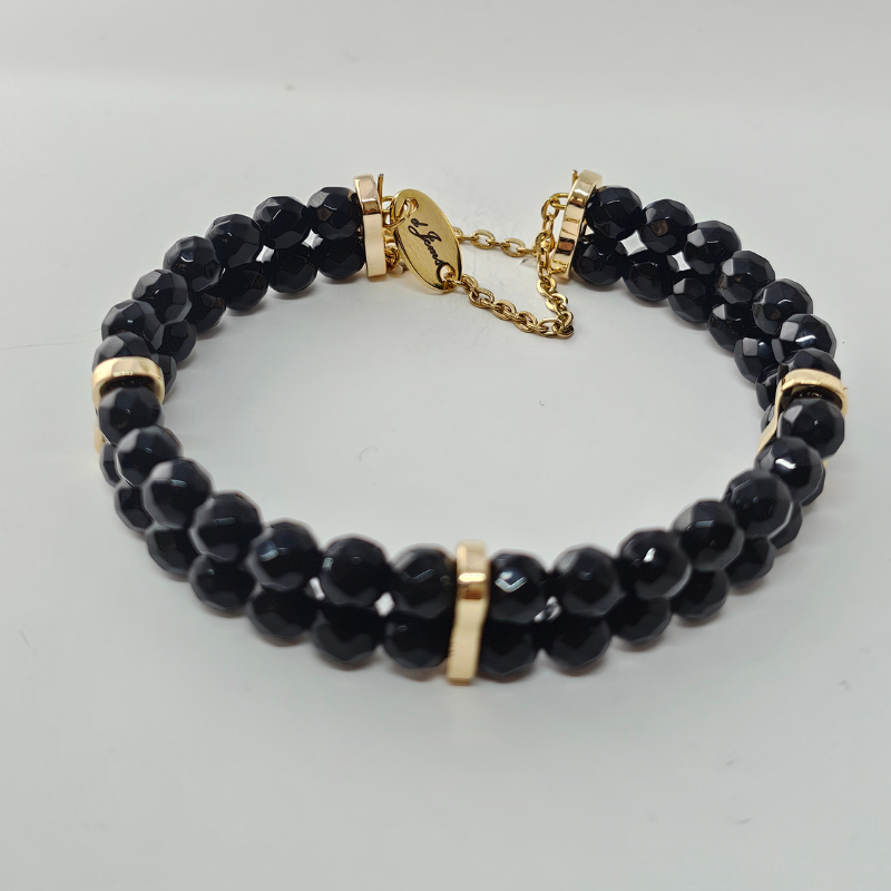 A double-strand black beaded bracelet with faceted round beads and gold-tone spacers and clasp. The bracelet features a delicate gold chain and an oval tag engraved with cursive text.