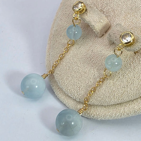 Pair of earrings with gold chains and light blue beads on a textured surface.