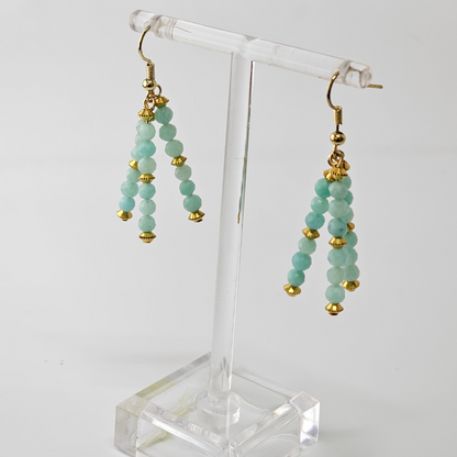 Teal beaded earrings with gold accents on a clear stand against a white background